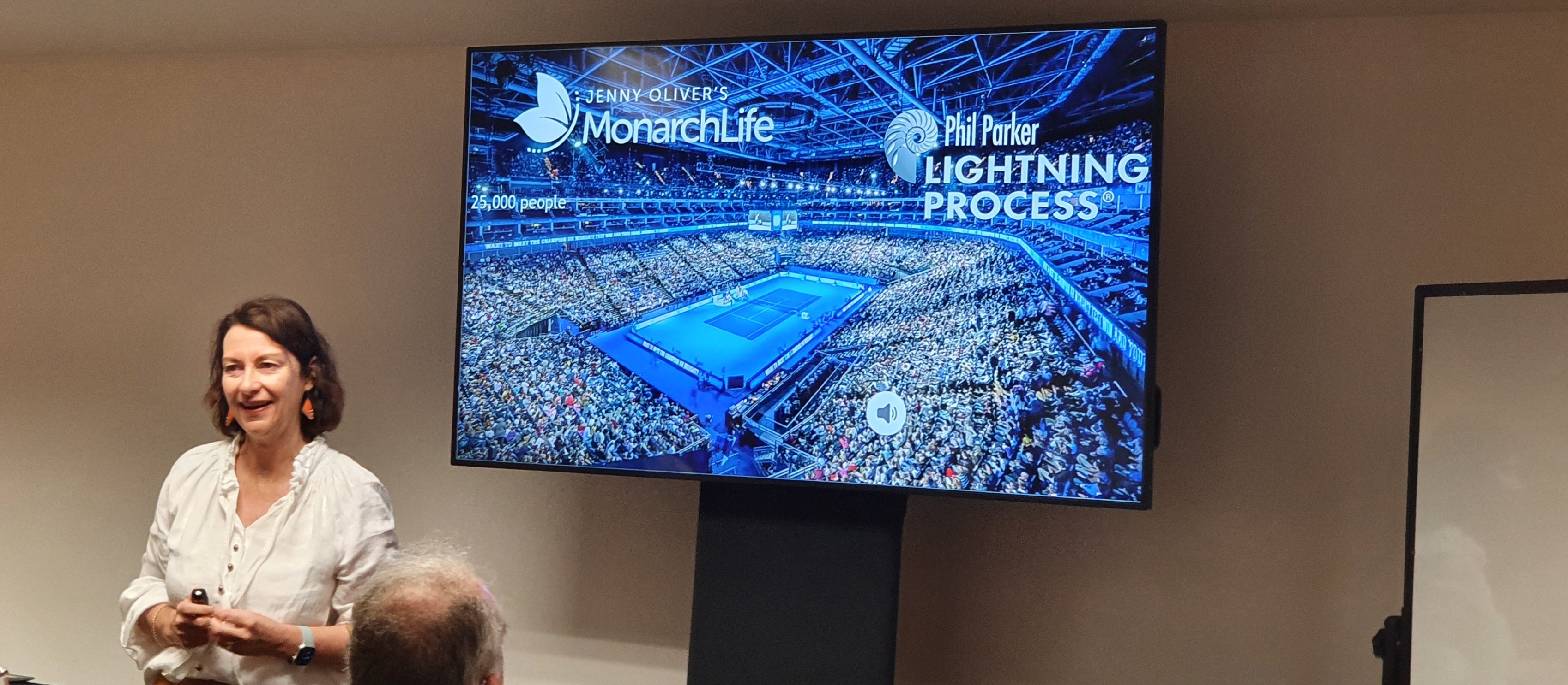 Image shows a woman, Jenny Oliver, standing beside a television screen during a presentation on the Lightning Process