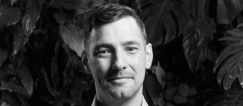 Black and white image shows man with his arms crossed, looking at the camera, with leafy trees in the background