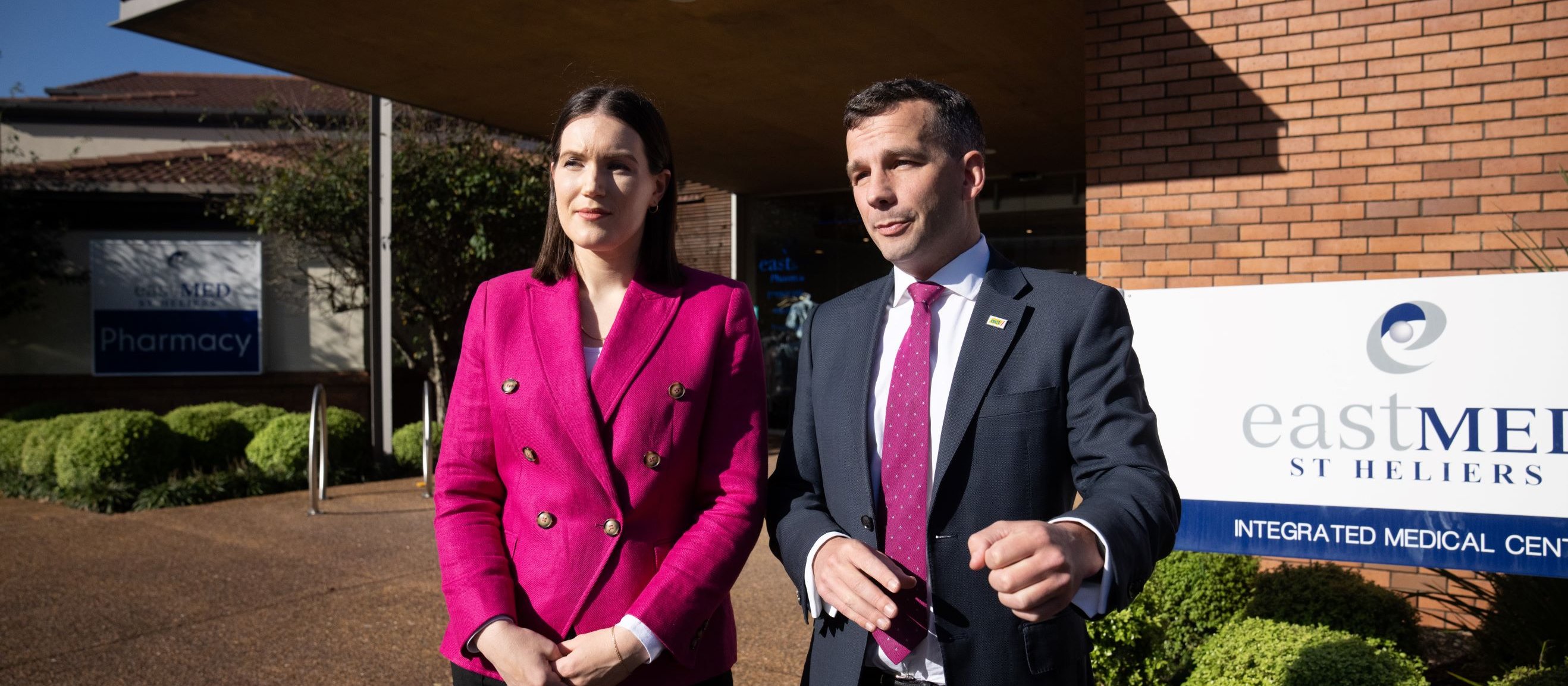 Image shows Brooke van Velden in bright pink blazer and black trousers standing beside David Seymour in a dark suit and pink tie, outside a redbrick medical centre