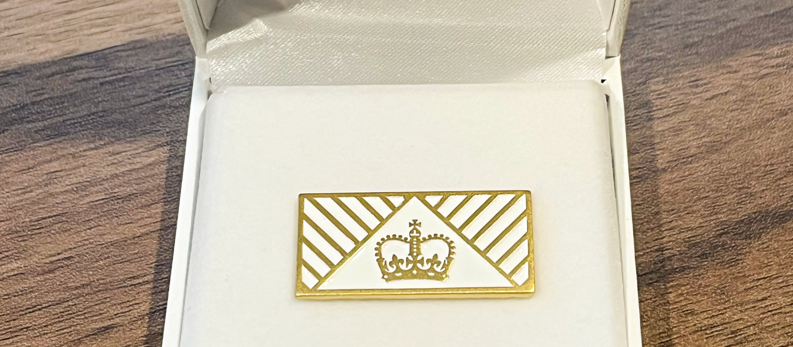 A gold lapel pin, with a crown in the centre, sits on a white display case with the NZ coat of arms on the underside of the lid
