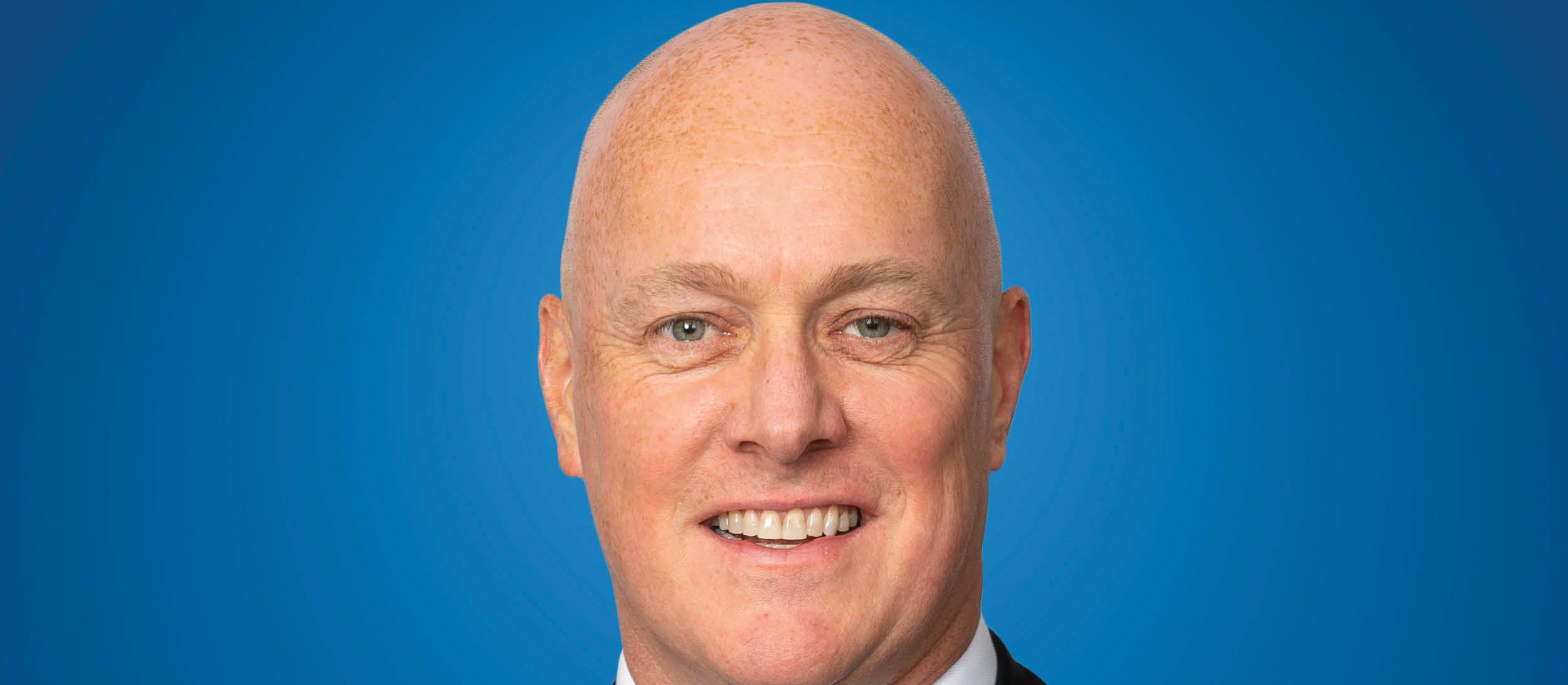 Image shows bald man in black suit, white shirt and blue tie, smiling at camera, against a blue background