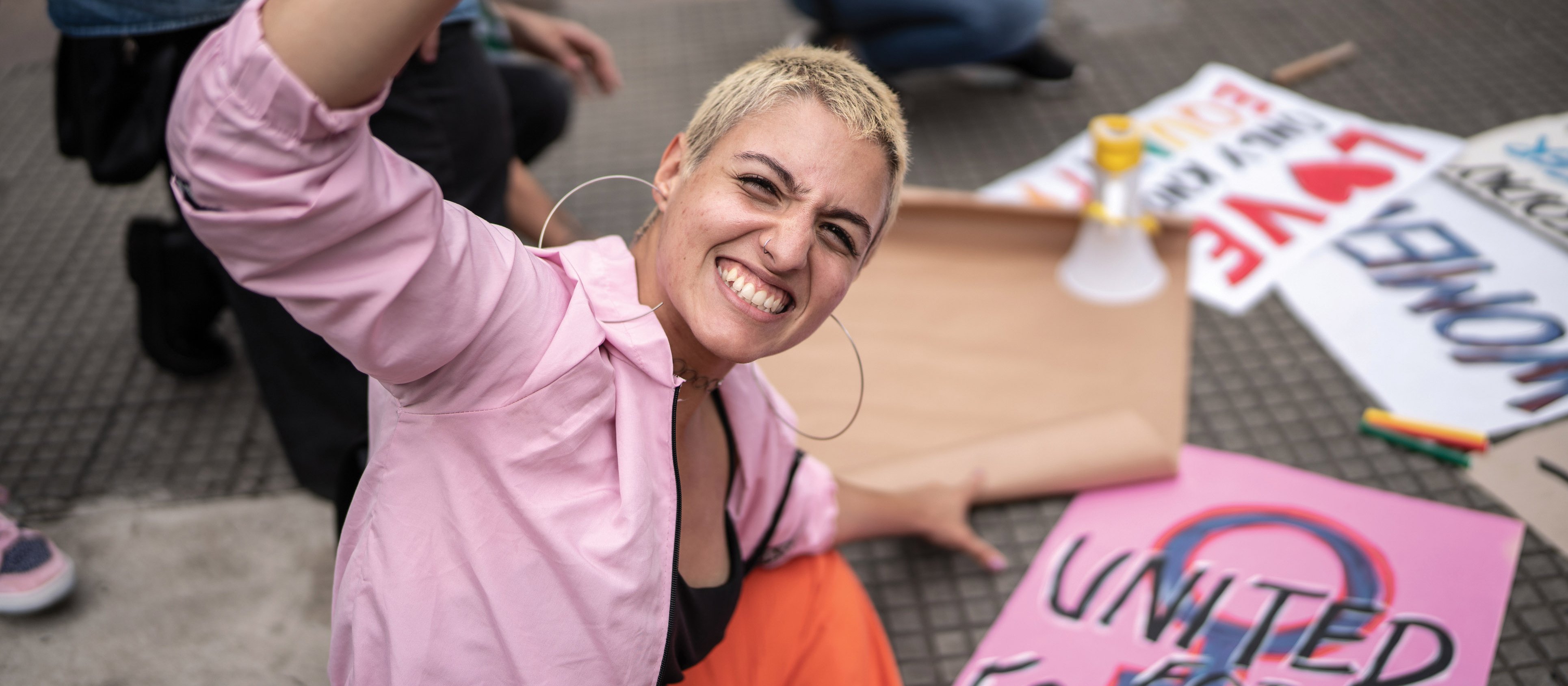 Young woman in a pink shirt and black top with cropped blonde hair raises a fist in the air as she kneels in front of protest signs, one reading 'united for equality'