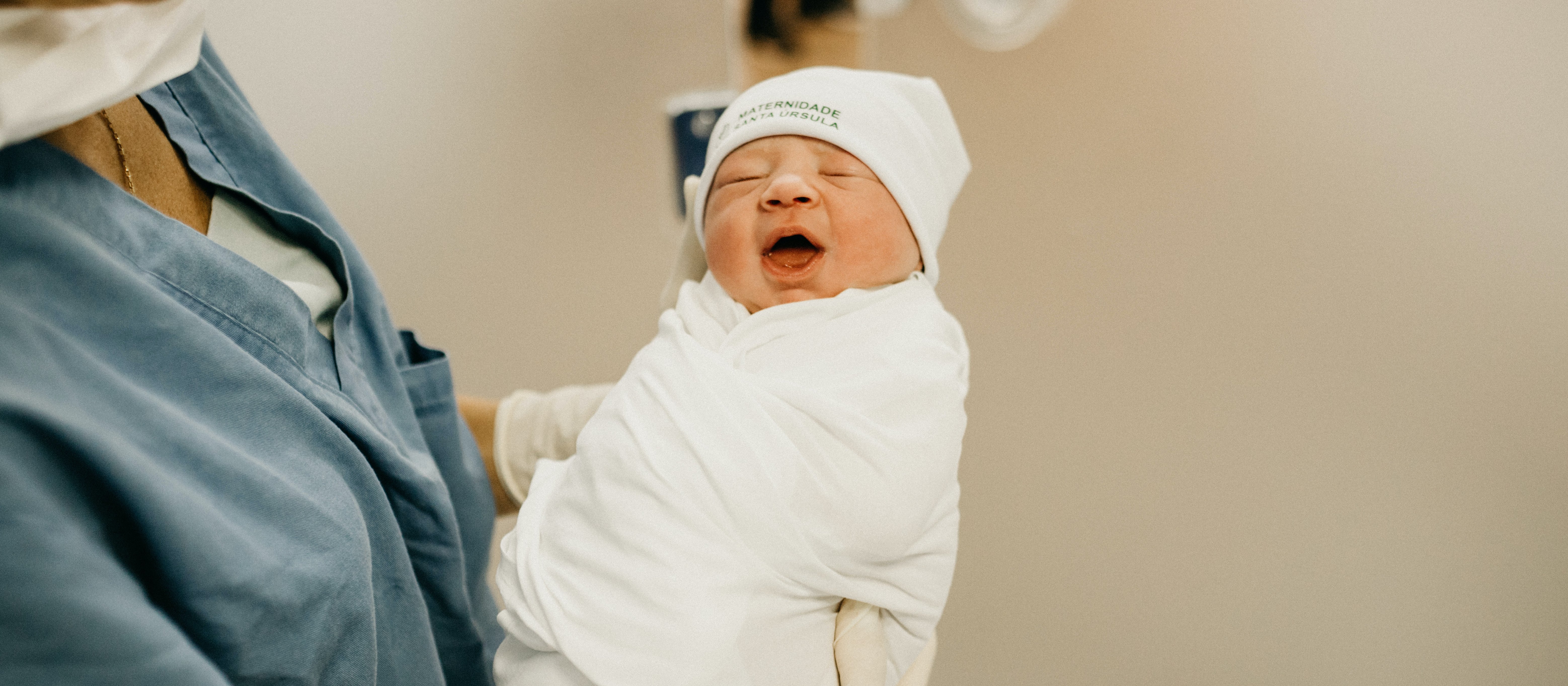 A newborn baby wrapped in a white swaddle and hat, being held by an adult in blue shirt, hospital room