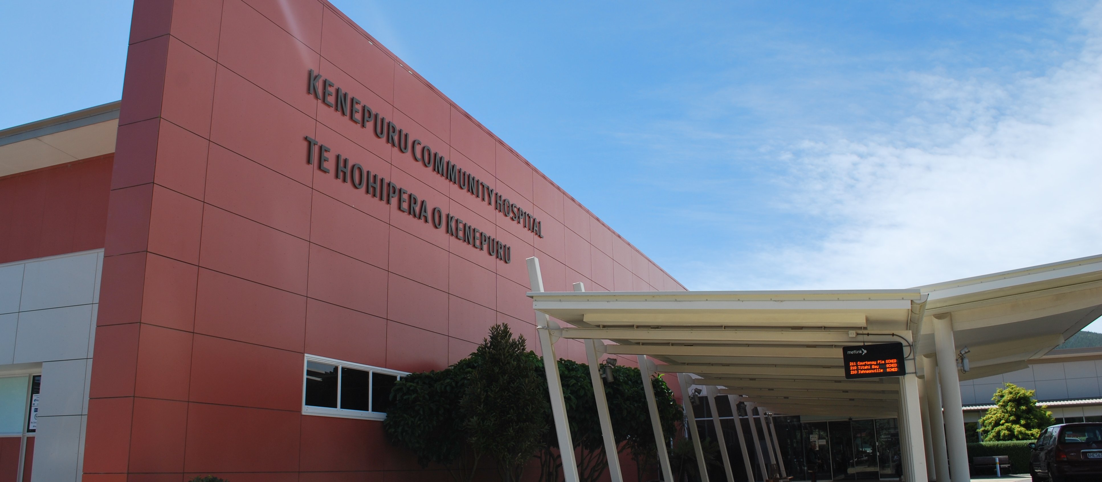 Side-view of Kenepuru Community Hospital, red-clad building with the name in large black writing on the front. White covered footpaths and a bus stop sign out the front