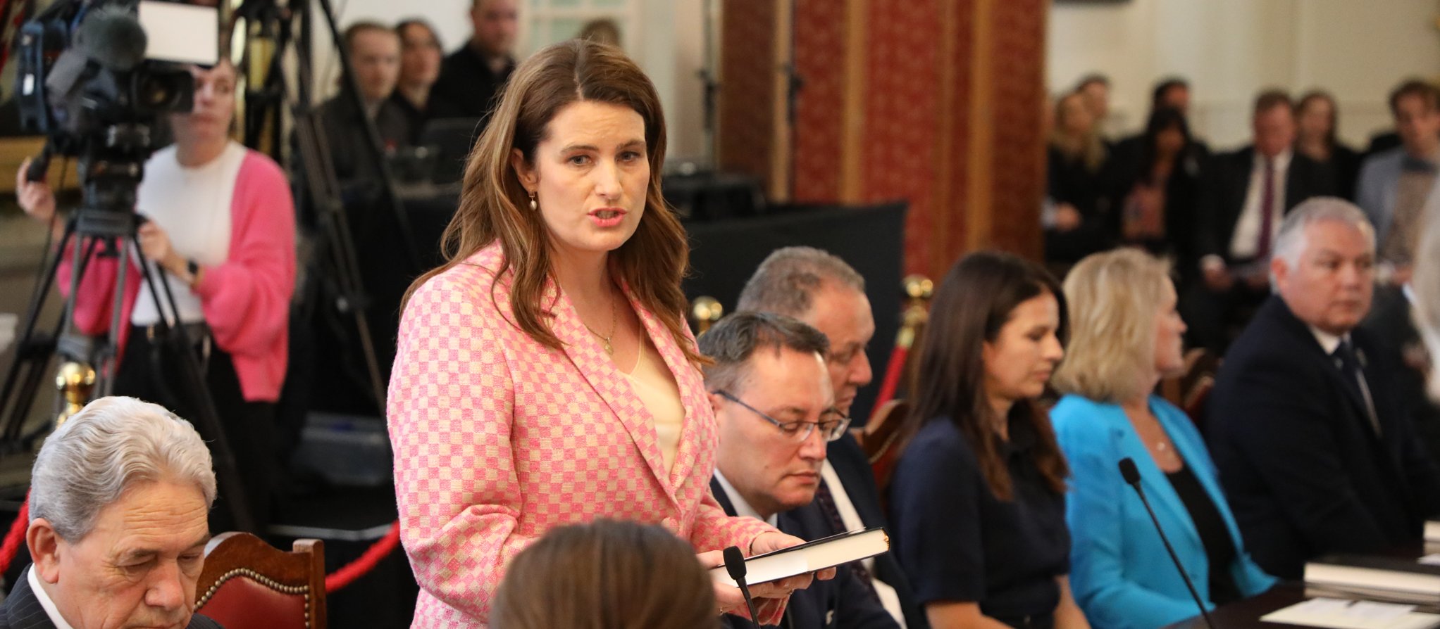 Woman with brown here and pink blazer standing up and speaking at a board table, surrounded by other people, media cameras in the background