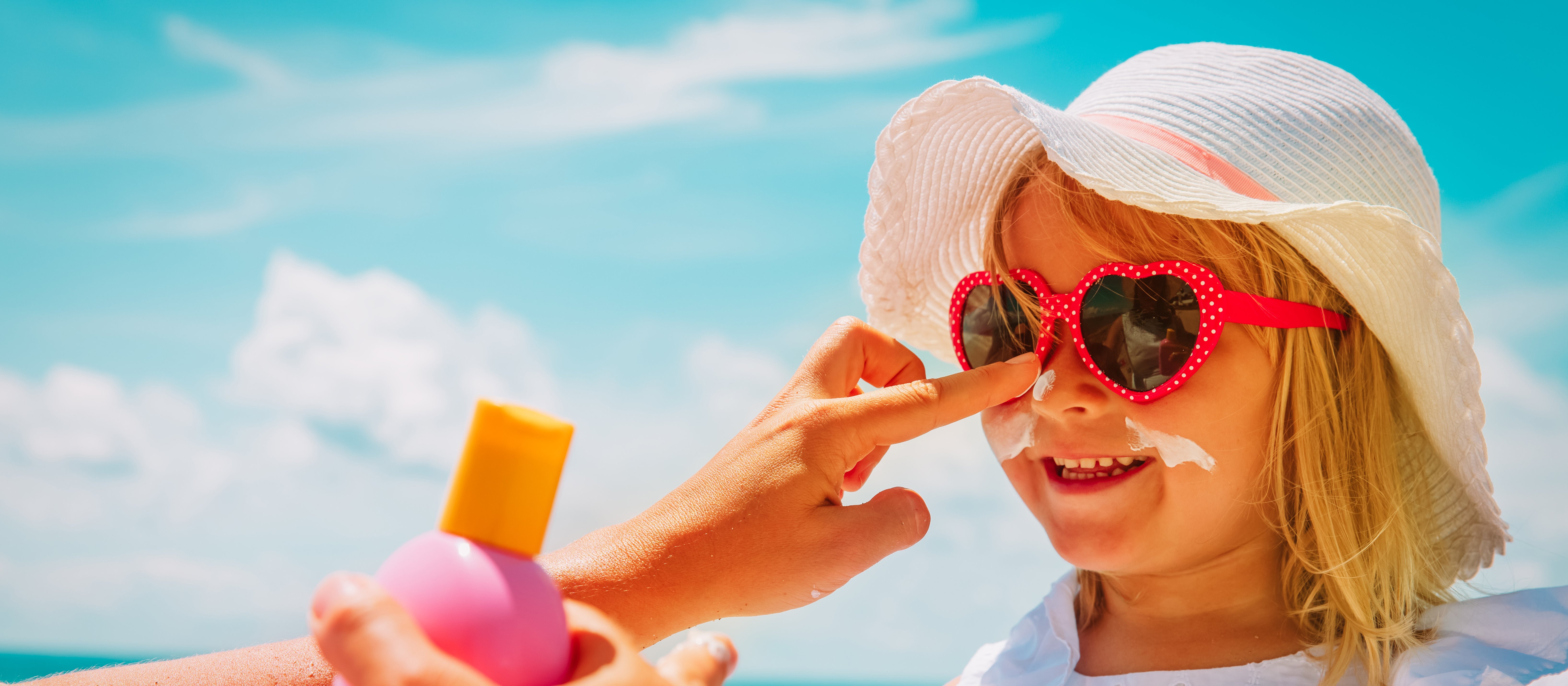 Blonde girl wearing white dress and sunhat, pink sunglasses, getting sunscreen applied to her face