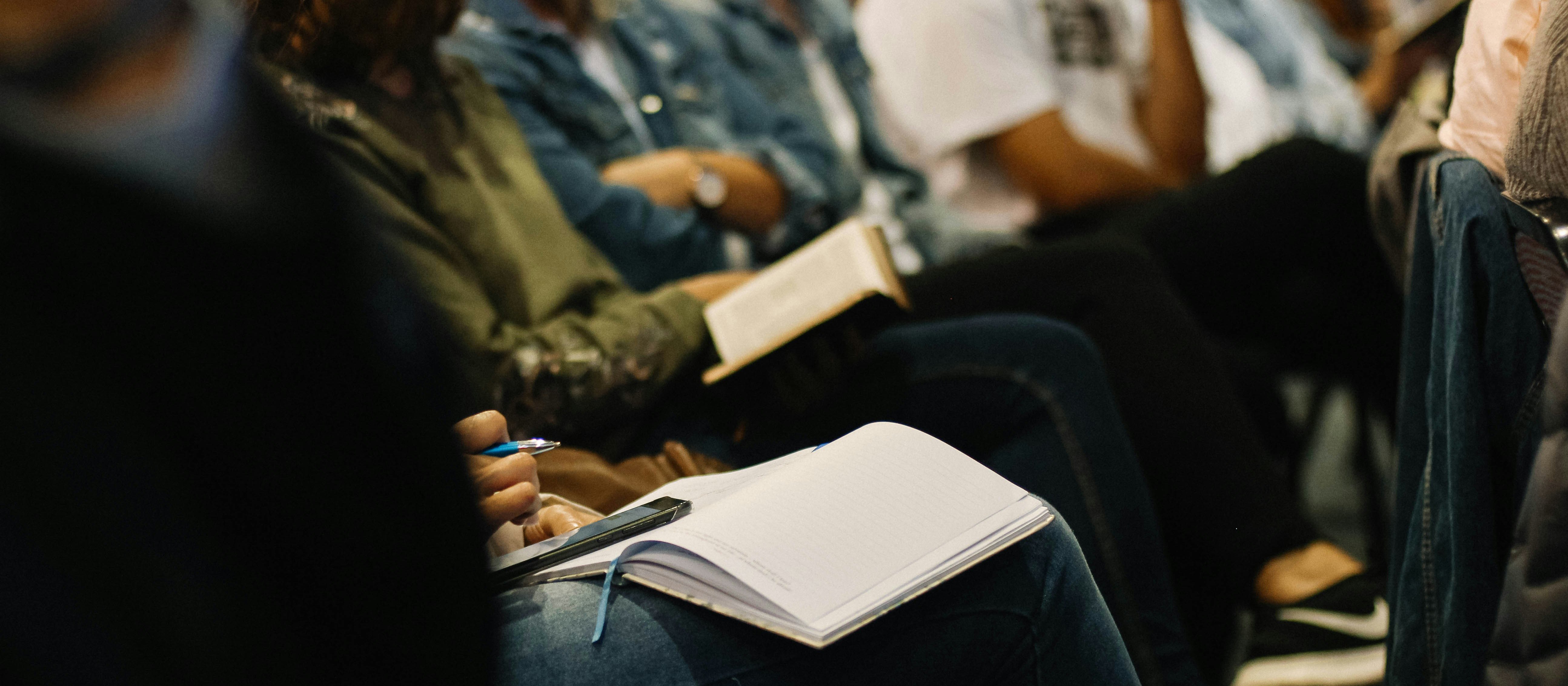 Image shows a row of seated people books and notepads on their knees, taking notes at a conference