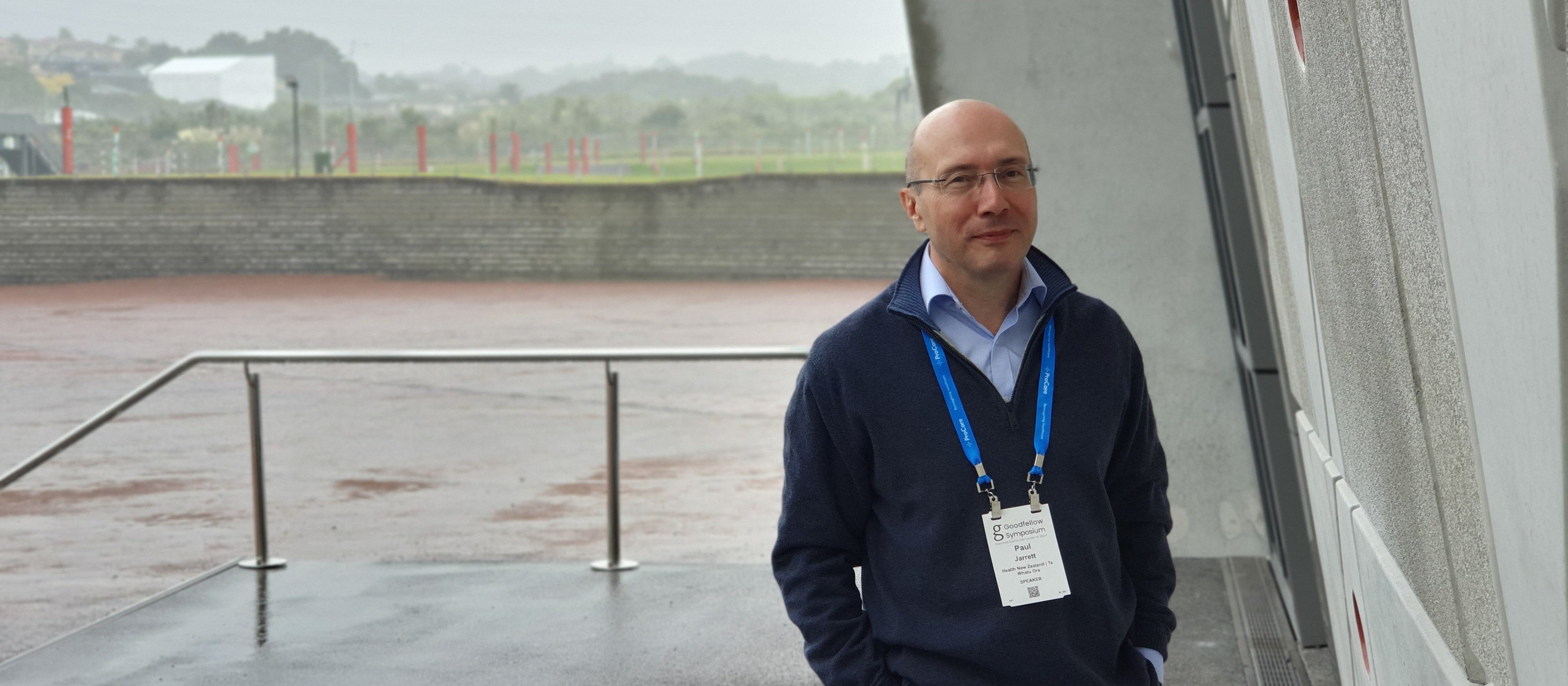 Image shows bald man in navy jumper standing outside conference centre, grey rainy background