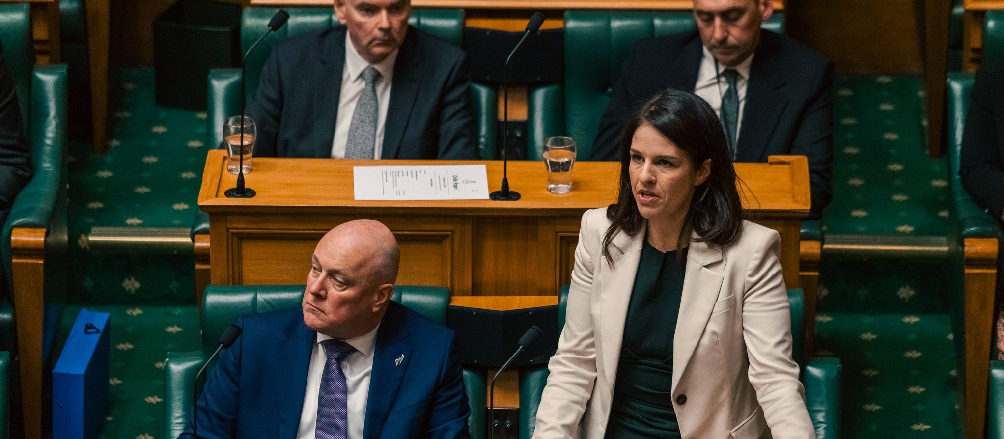 Minister Erica Stanford stands while speaking in Parliament, Prime Minister Christopher Luxon sits beside her
