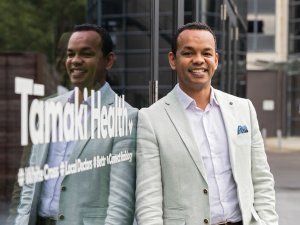 Image shows man in light blazer and white shirt leaning against a reflective window with the sign Tāmaki Health in white writing