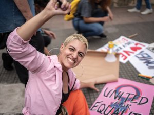 Young woman in a pink shirt and black top with cropped blonde hair raises a fist in the air as she kneels in front of protest signs, one reading 'united for equality'
