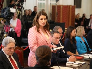 Woman with brown here and pink blazer standing up and speaking at a board table, surrounded by other people, media cameras in the background