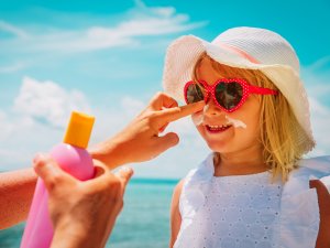 Blonde girl wearing white dress and sunhat, pink sunglasses, getting sunscreen applied to her face