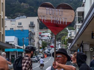 Image shows a man in a black hat and sunglasses holding a heart-shaped sign with black, white and red stripes, which reads 'Toitū te tiriti'
