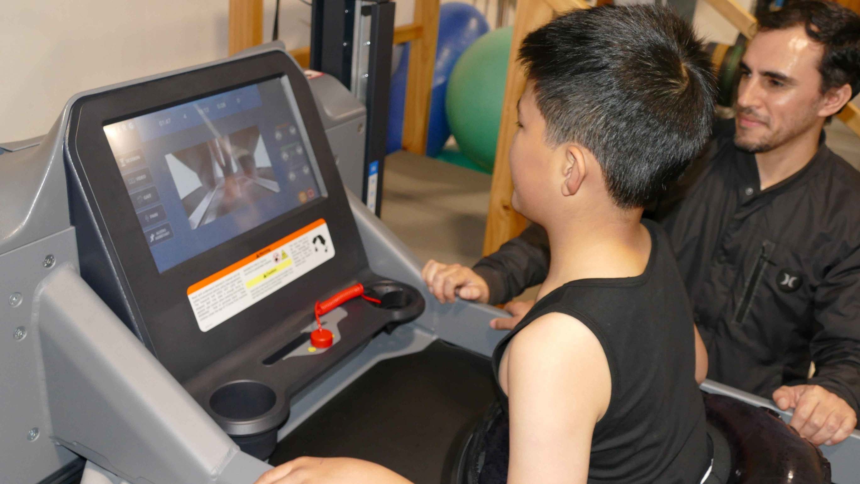 Image shows boy with dark hair wearing a singlet on a treadmill-like machine looking at a screen with a male researcher with dark hair and a black shirt looking on