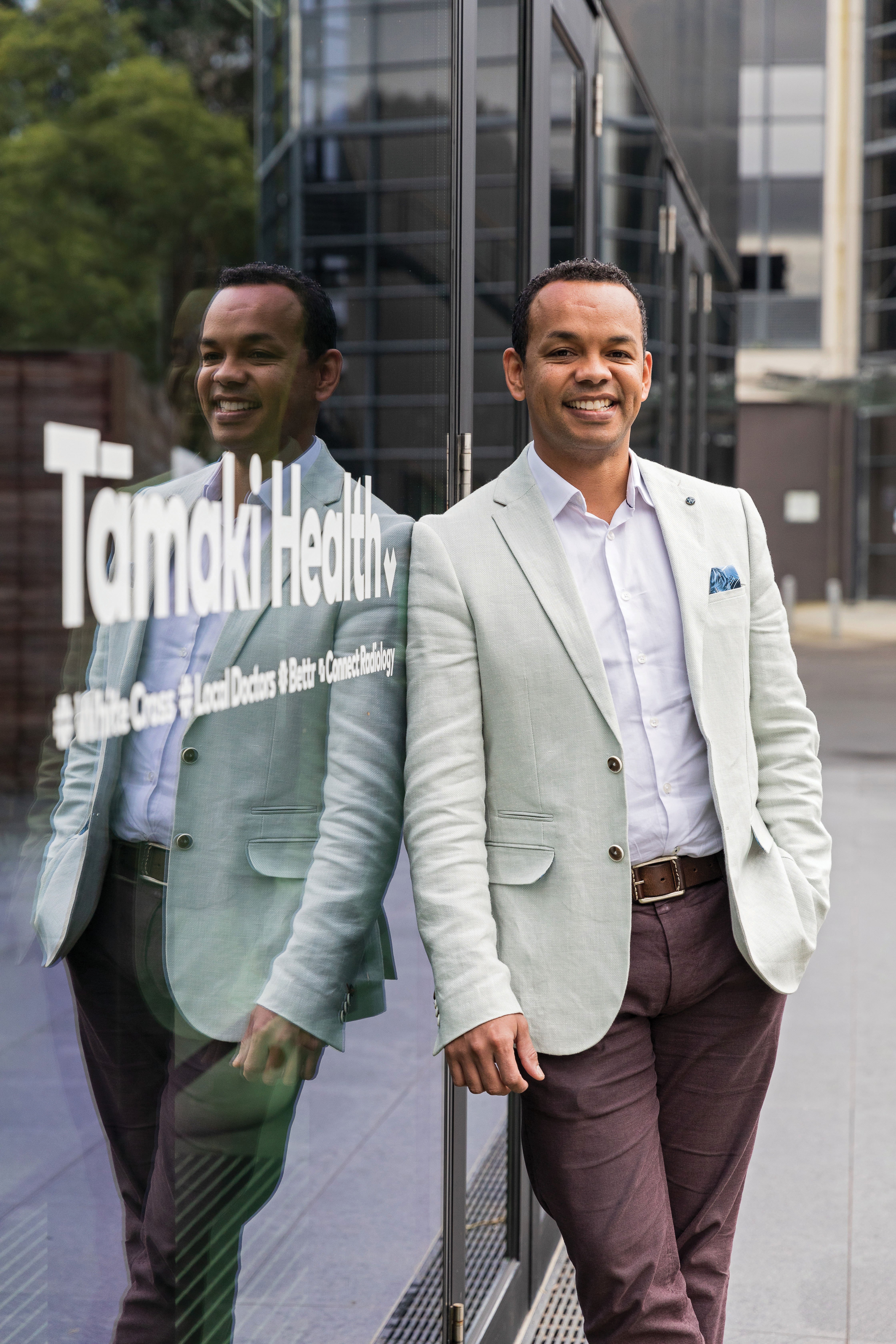 Image shows man in light blazer and white shirt leaning against a reflective window with the sign Tāmaki Health in white writing