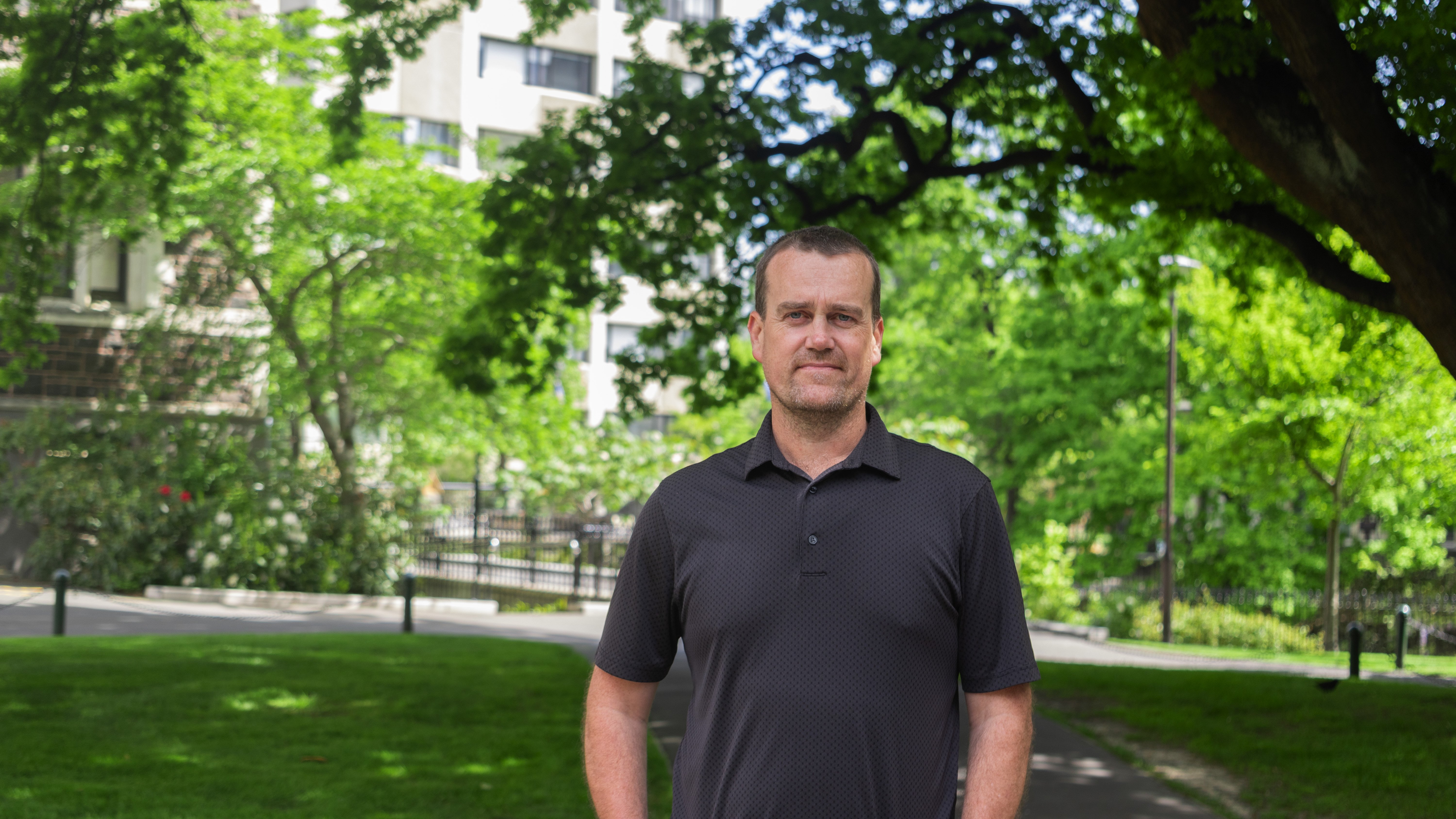 Man in dark polo t-shirt standing outside with gardens and trees in background